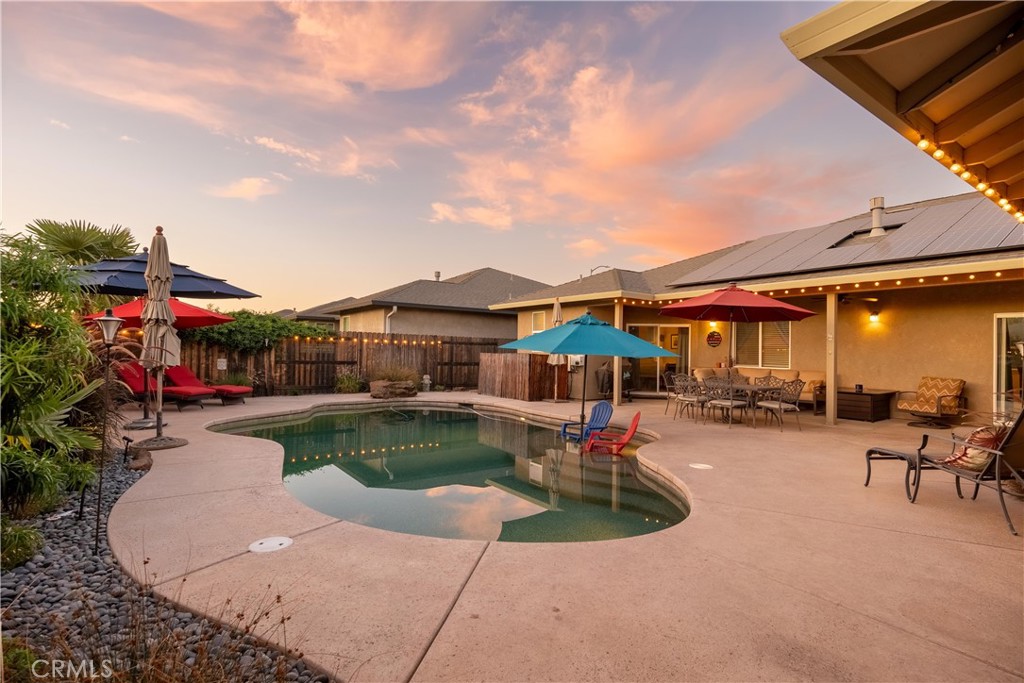 Backyard pool beside a furnished covered patio, with solar panels and string lights at sunset.