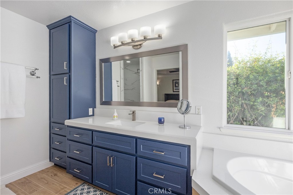 Bathroom vanity with blue cabinets, a wide mirror, and a window beside the bathtub.