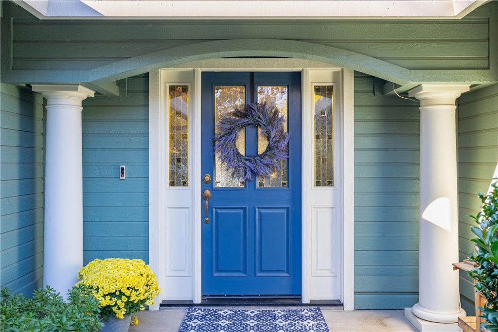 Blue double front door with a purple wreath, white columns, yellow flowers, and patterned doormat.