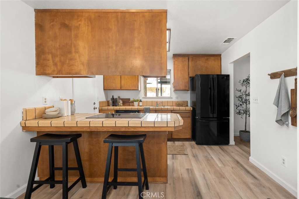 Kitchen with warm wood cabinets, tiled breakfast bar, black refrigerator, and two stools.