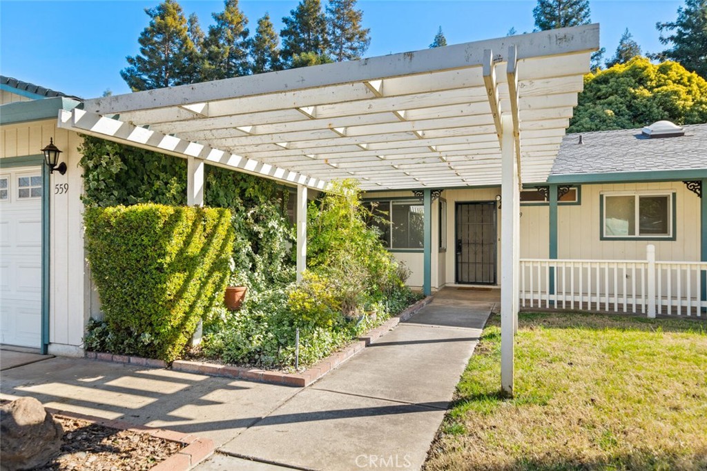 Home entrance beneath a white pergola, with a trimmed hedge and garden bed along the walkway.