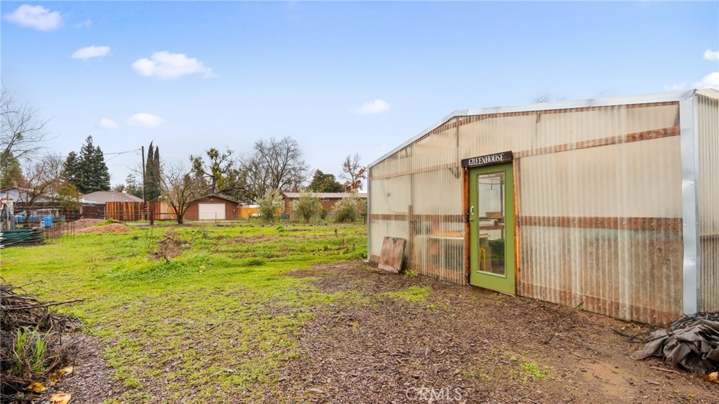Backyard greenhouse with a green door, beside garden beds and neighboring outbuildings.