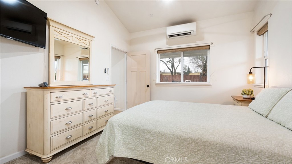 Furnished bedroom with a large bed, light wood dresser, wall-mounted television, and window overlooking a fenced yard.