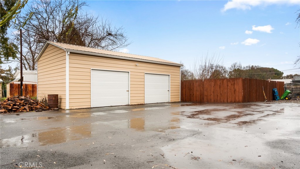 Tan detached garage with two white roll-up doors beside a wet paved driveway and wooden fence.