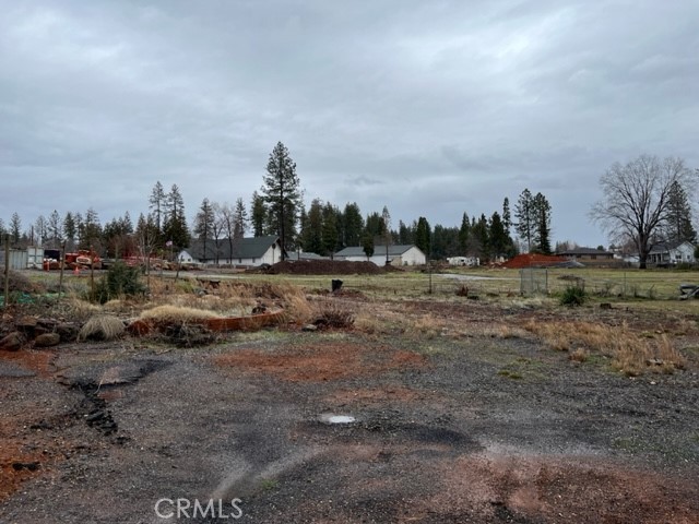 Vacant lot with patchy grass and gravel, neighboring homes and trees in the distance.