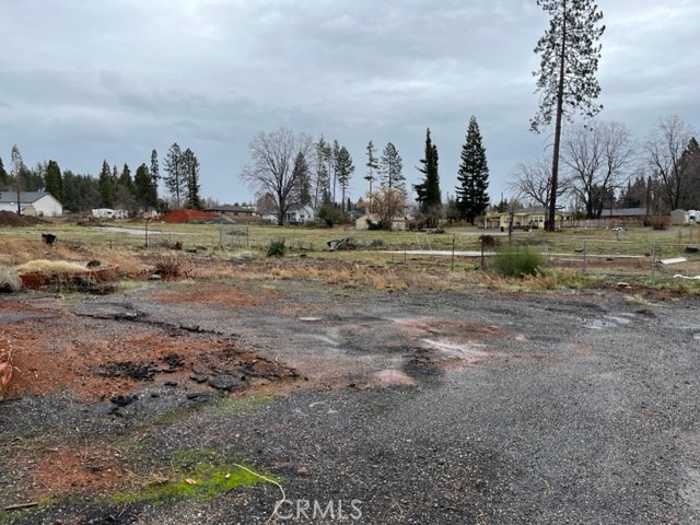 Vacant lot with rough gravel ground, patchy grass, and trees and homes in the distance.