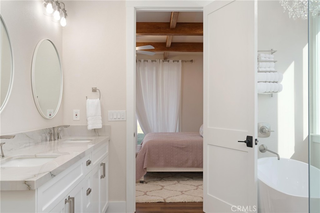 Bathroom vanity and tub frame a doorway into a bedroom with exposed ceiling beams.