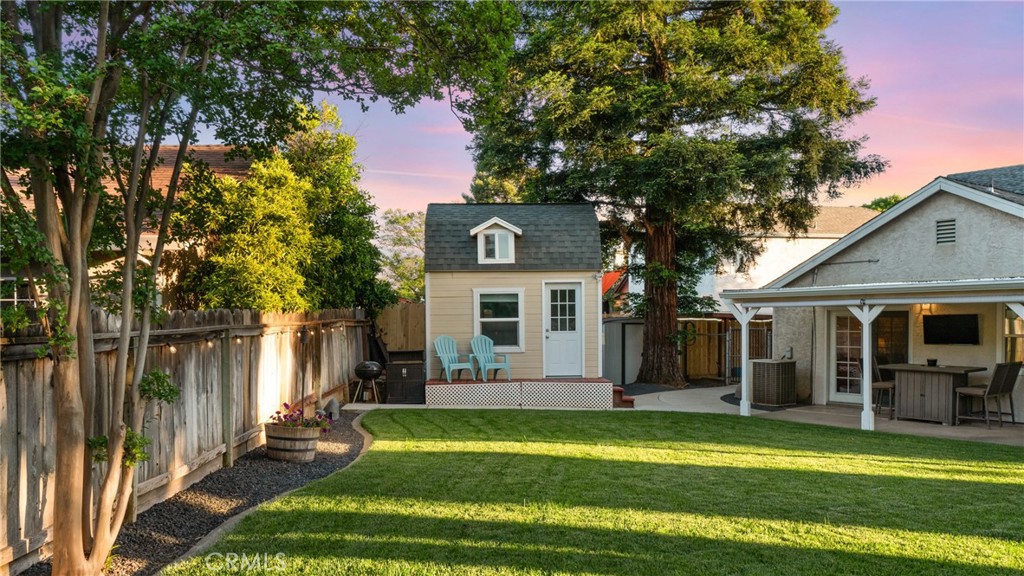 Backyard with a detached cottage, blue chairs, mature trees, and a covered patio beside the lawn.