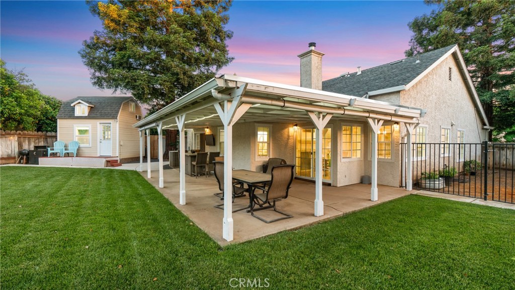 House with a furnished covered patio, broad lawn, and small detached outbuilding at dusk.