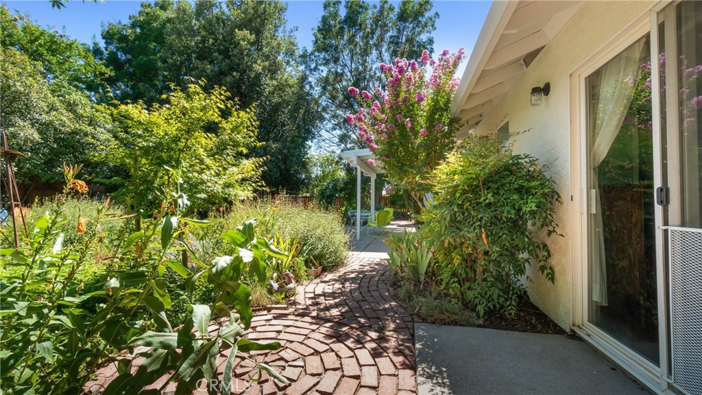 Garden patio with a curved brick path, flowering plants, and a covered seating area.