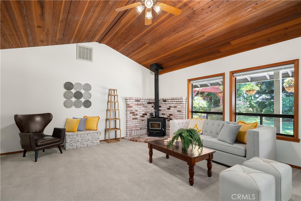 Living room with a vaulted wood ceiling, brick fireplace, black wood stove, and large windows.