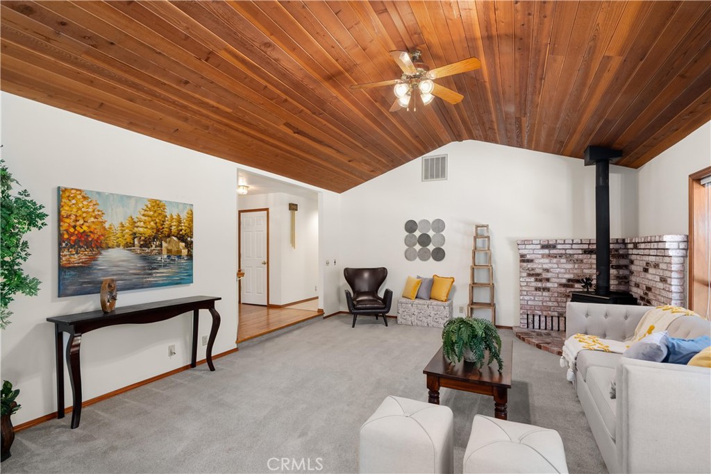 Living room with vaulted wood ceiling, gray carpet, and brick fireplace with a black wood stove.