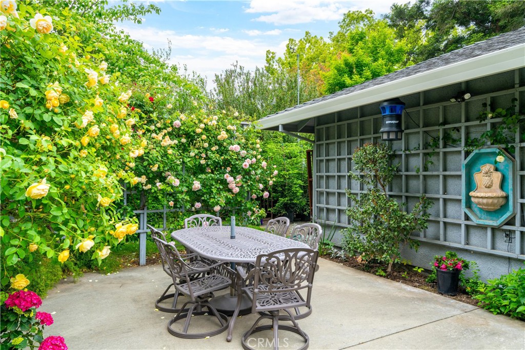 Patio dining area with metal table and chairs, flowering roses, and a wall-mounted fountain.