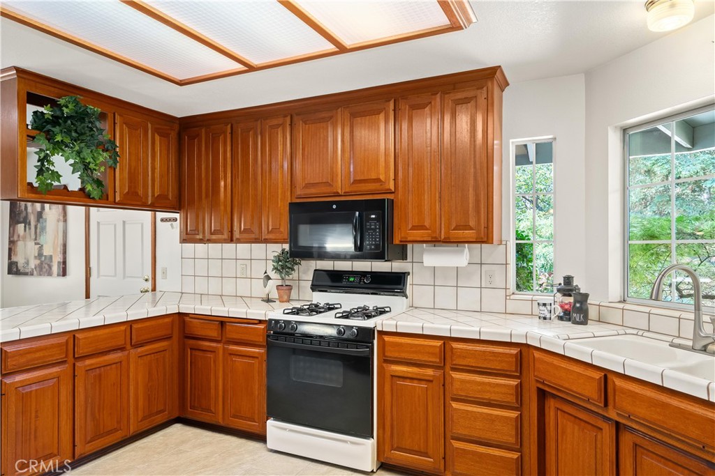 Kitchen with warm wood cabinets, tiled counters, gas range, and a sink beside a large garden-facing window.