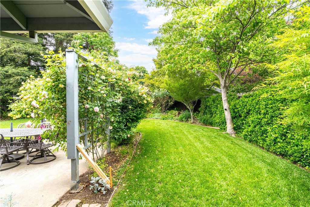 Landscaped backyard with a green lawn, flowering shrubs, mature trees, and a covered patio with outdoor furniture.