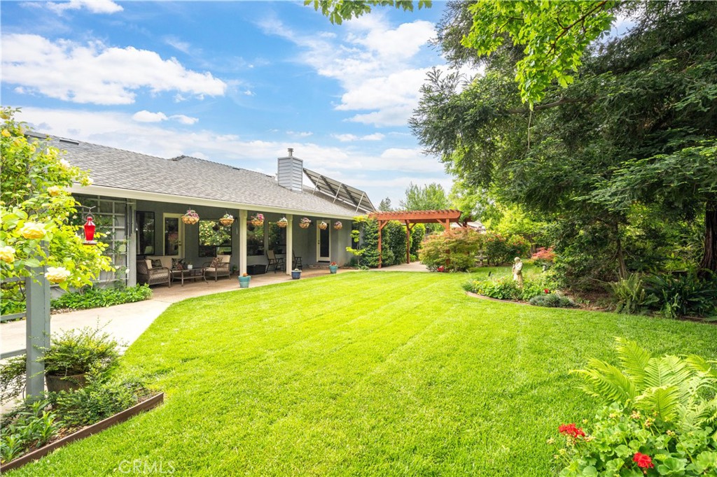 Home’s rear patio and broad lawn, framed by a wooden pergola, mature trees, and garden beds.