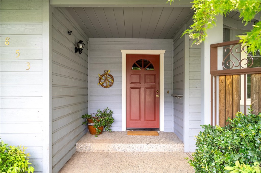 Covered front entry with a red paneled door, gray siding, and leafy plants.