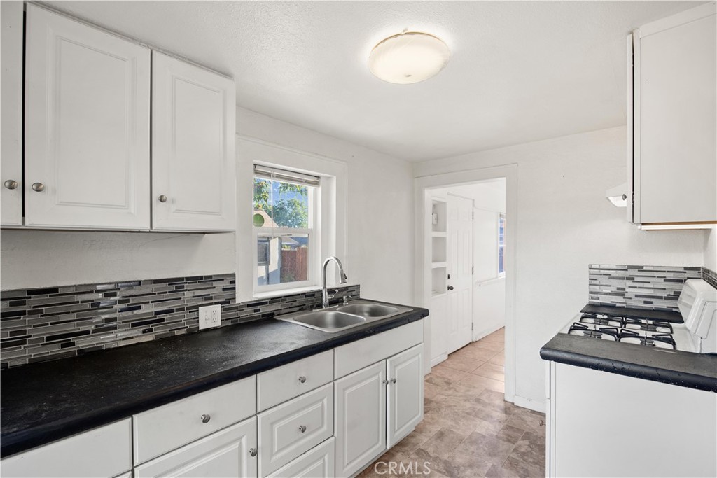 Vacant kitchen with white cabinets, dark counters, tile backsplash, double sink, and gas range.