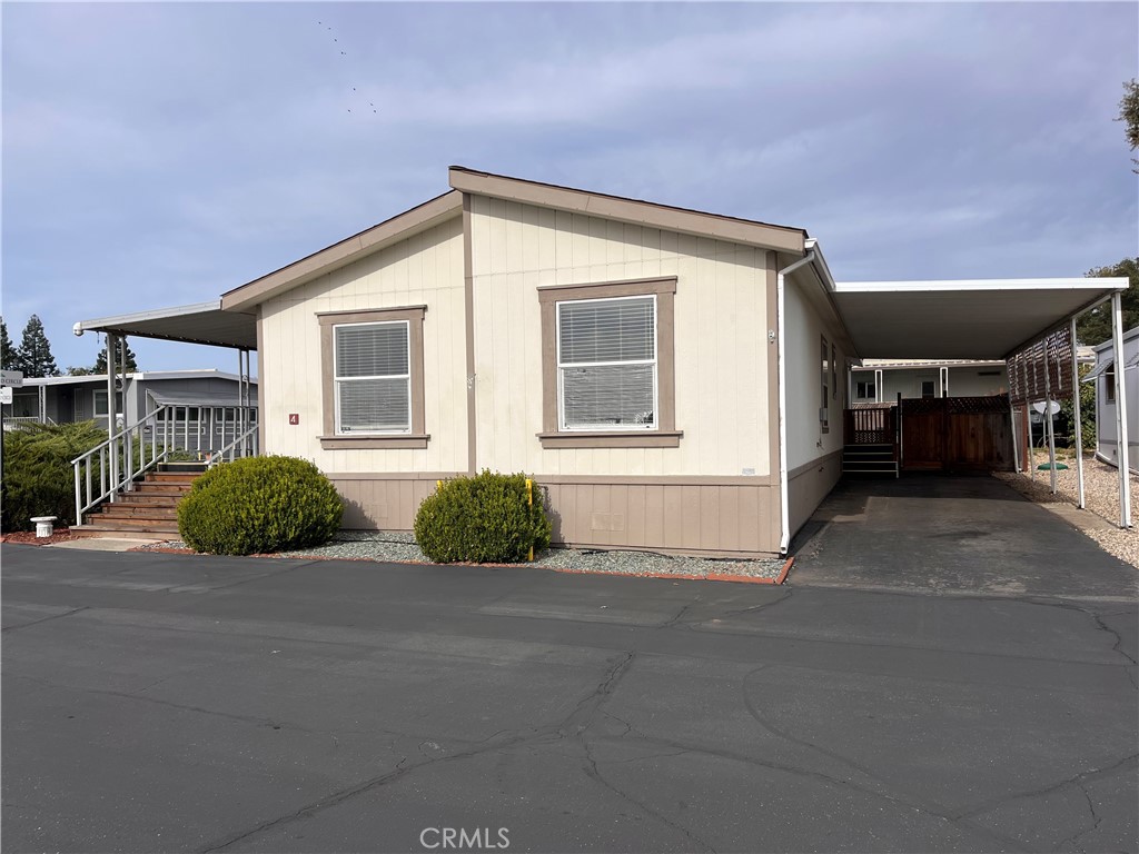 Beige manufactured home with a raised front entry, shrubs, and a covered carport along the right side.