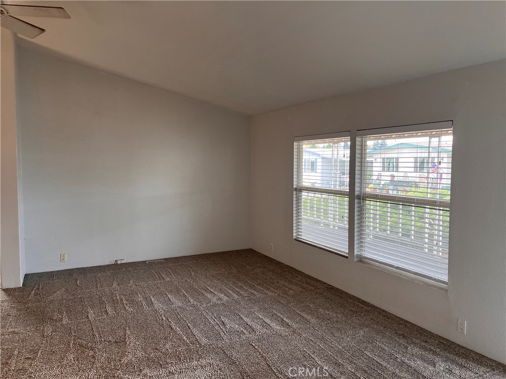 Vacant living room with brown carpet and two large windows fitted with blinds.