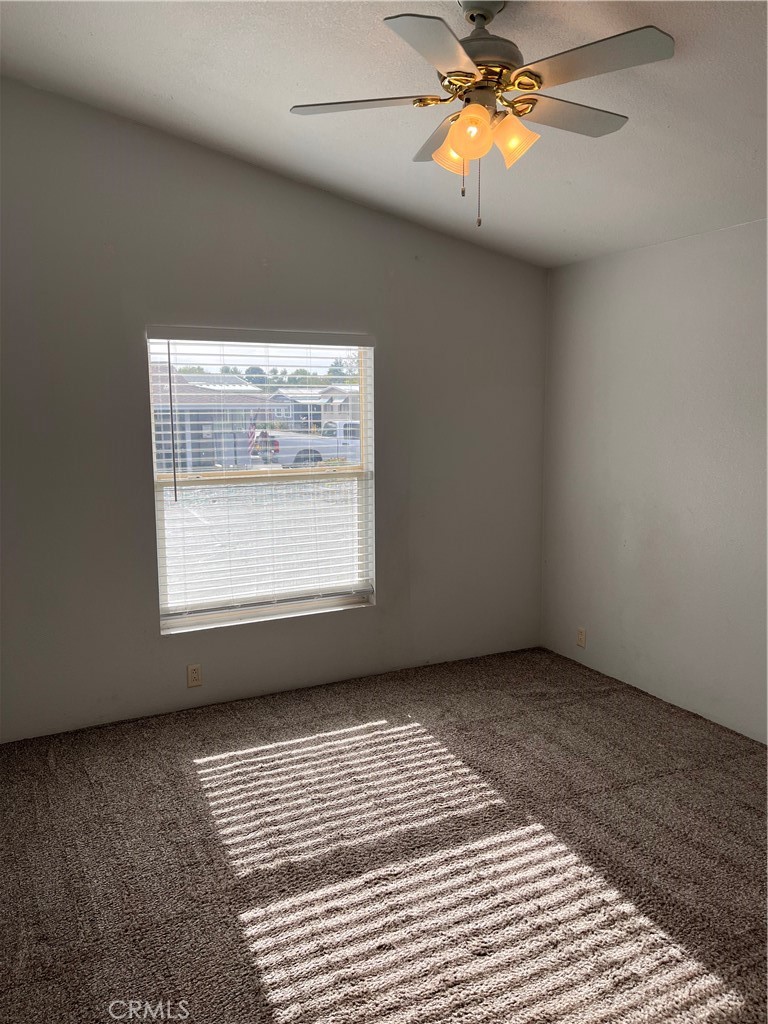 Empty carpeted room with a sloped ceiling, ceiling fan, and sunlight through window blinds.