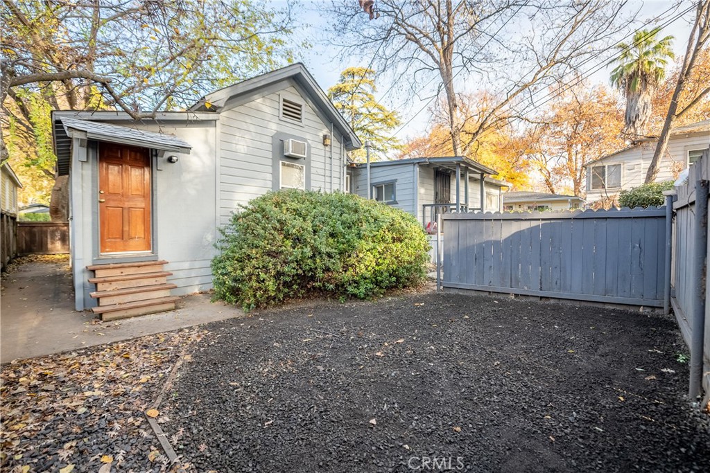 Gray house with a wooden entry door beside a fenced gravel yard and shrubs.