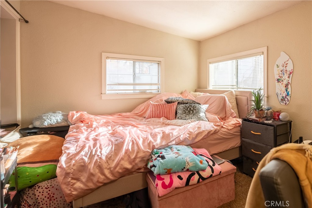 Furnished bedroom with rumpled pink bedding, two windows with blinds, and a dark bedside table.