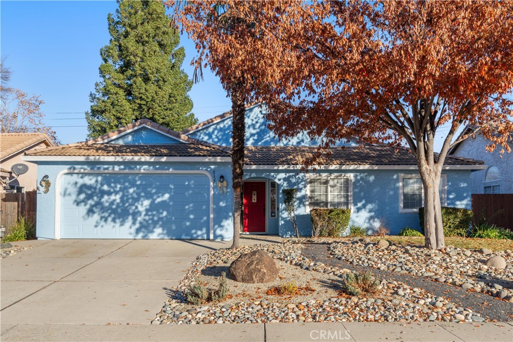 Blue single-story home with a red front door, two-car garage, and rock landscaping beneath autumn trees.