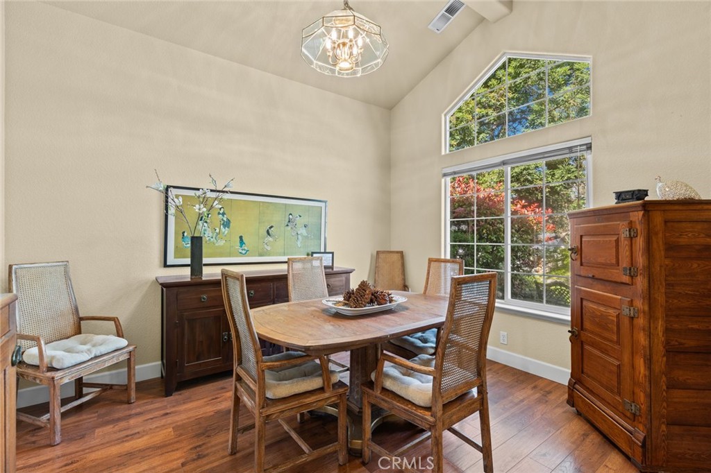 Dining room with a wood table, woven chairs, vaulted ceiling, and tall windows overlooking trees.