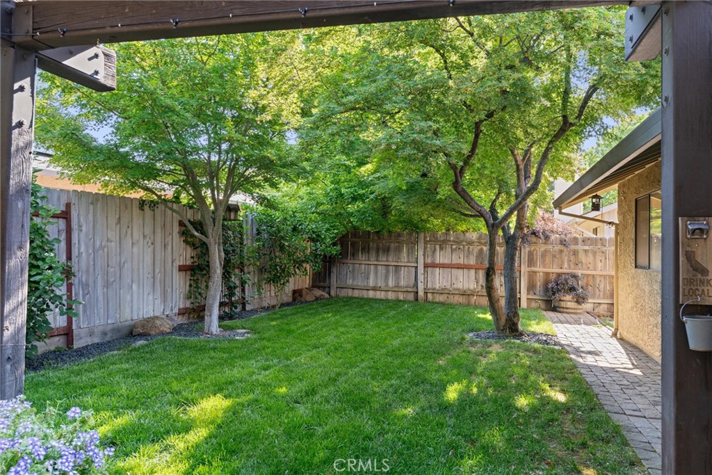Fenced backyard with a broad lawn, mature trees, and a paved walkway beside the house.