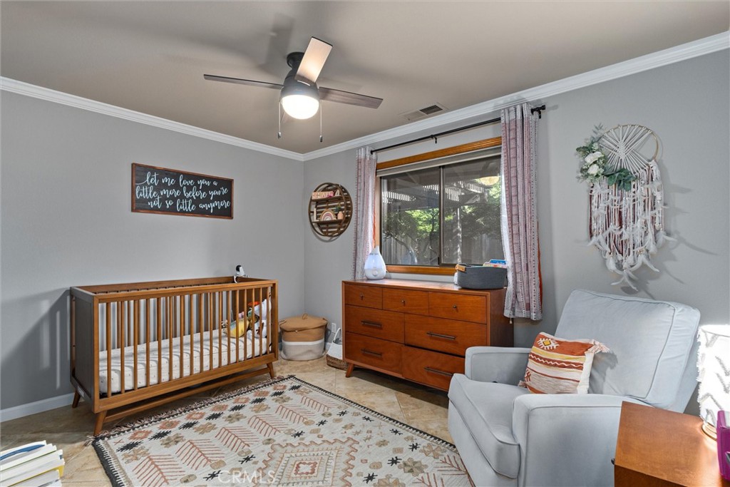Nursery with a wooden crib, matching dresser, gray armchair, patterned rug, and large curtained window.