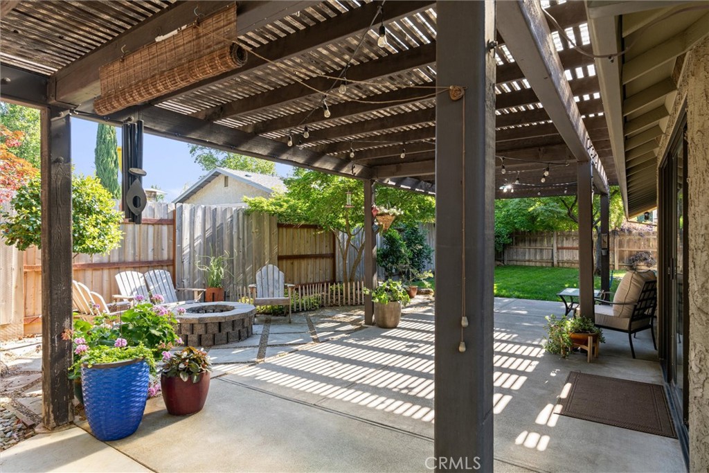 Covered backyard patio with a wooden pergola, potted flowers, and a stone fire pit.