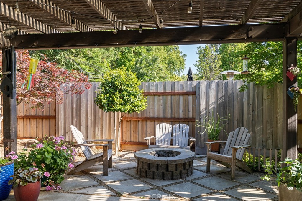 Fenced backyard patio under a wooden pergola, with Adirondack chairs around a circular stone fire pit.