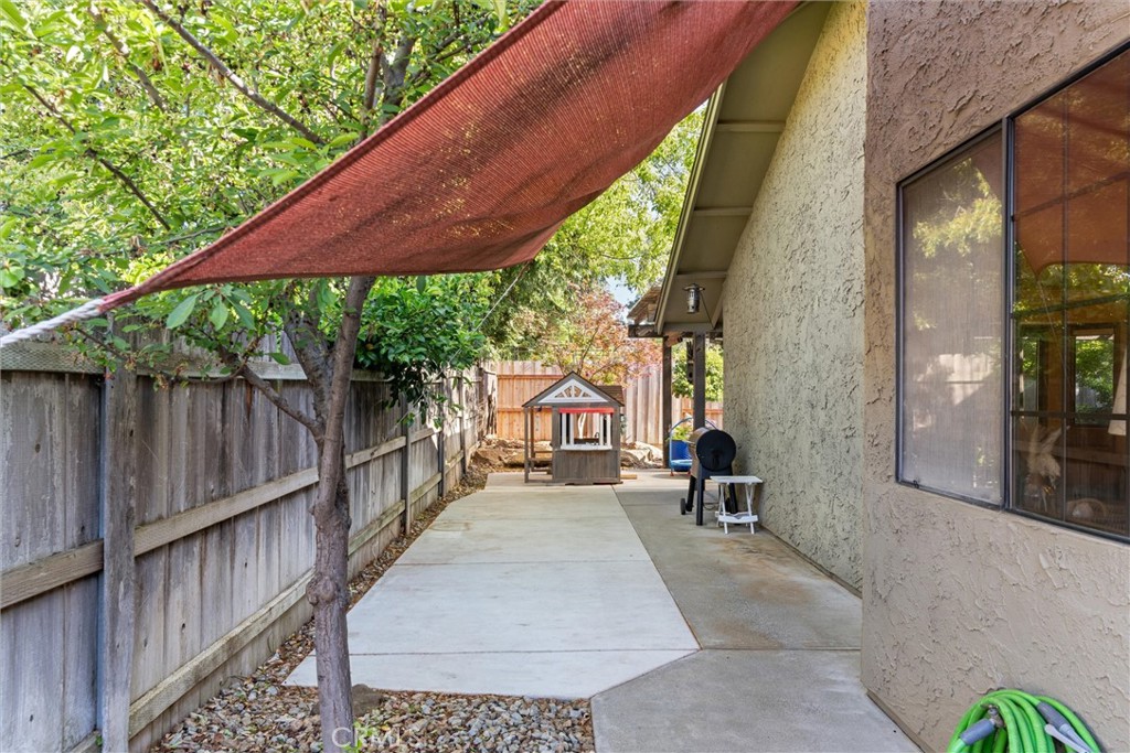 Side yard patio with a red shade sail, concrete walkway, and small playhouse beside a stucco home.