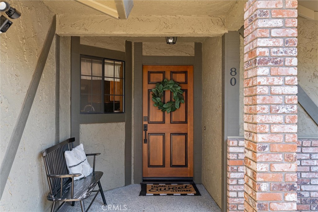Covered front porch with a wooden entry door, green wreath, black bench, and brick column.