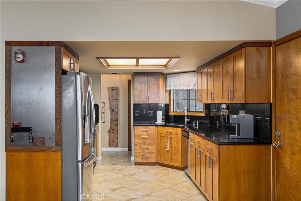 Kitchen with honey-toned wood cabinets, black countertops, stainless steel refrigerator, and a large ceiling skylight.
