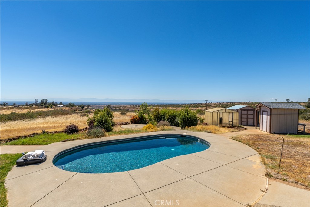Swimming pool and concrete deck beside sheds, with open fields and distant hills.