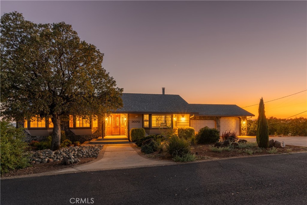 Single-story house at dusk with warm glowing windows, stone accents, and a large front-yard tree.