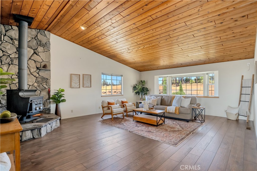 Staged living room with a vaulted wood ceiling, stone fireplace, and large windows overlooking open land.