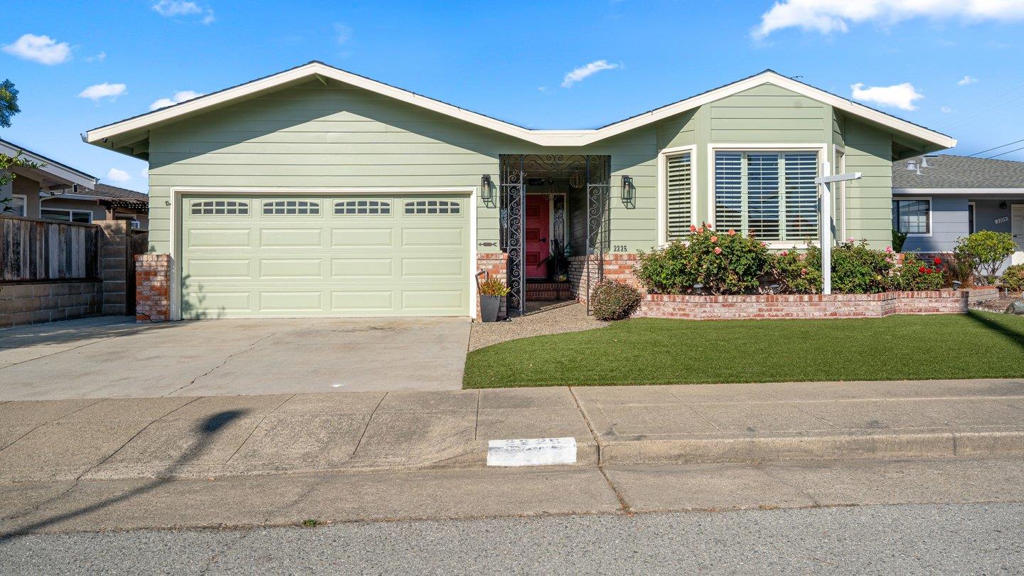 Pale green single-story home with a two-car garage, red entry door, and brick-edged lawn.