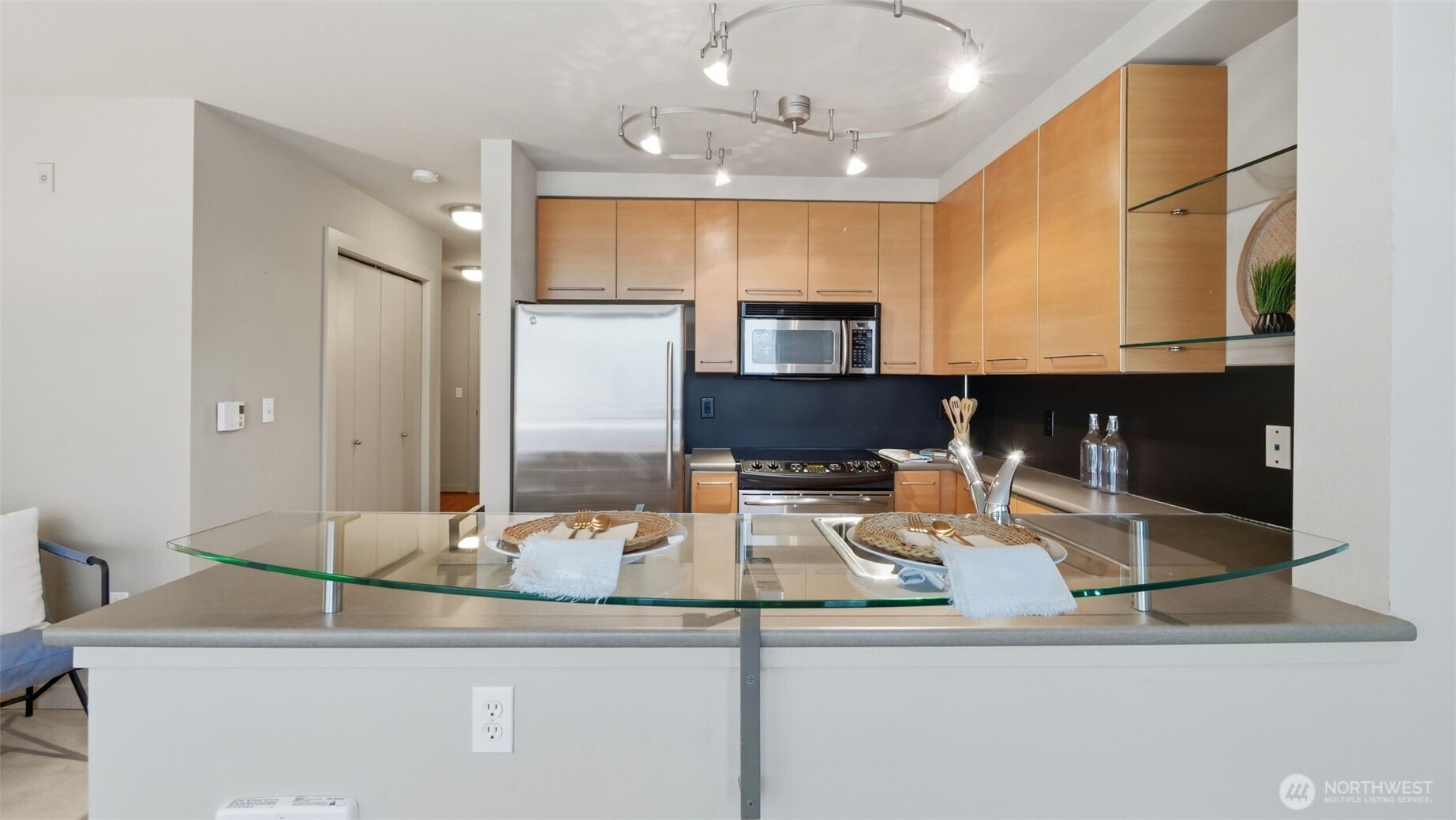 Condo kitchen with light wood cabinets, stainless steel appliances, and a glass counter set for two.