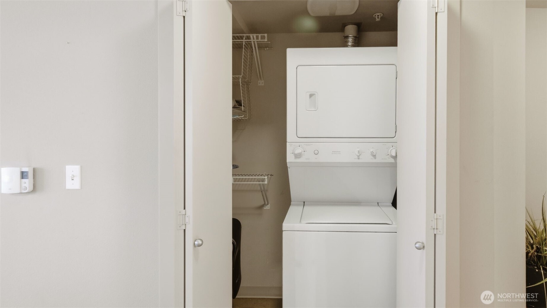 Laundry closet with a stacked white washer and dryer beside wire shelving.