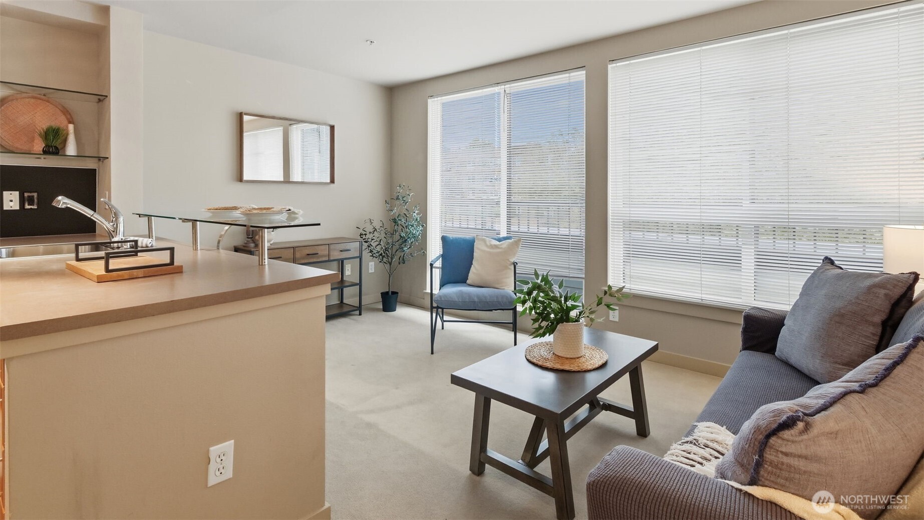 Furnished condo living room with a gray sofa, blue chair, and large windows with blinds.