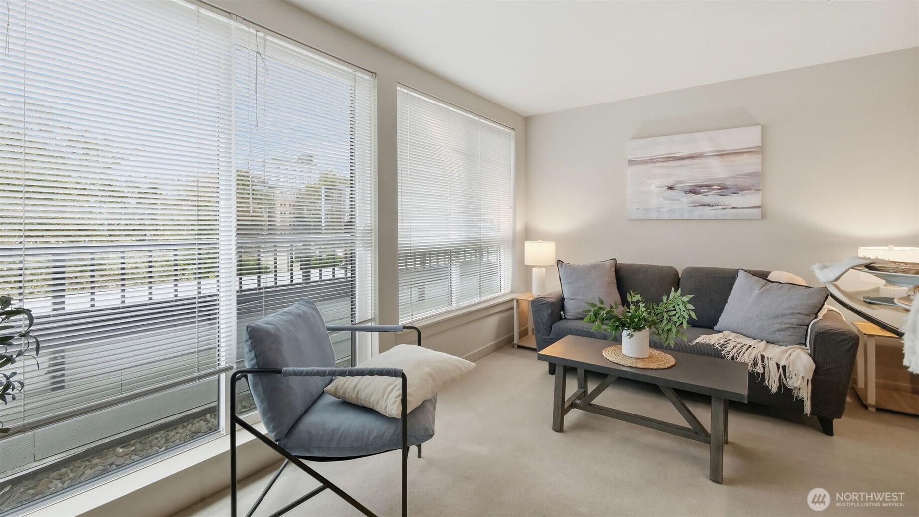 Staged living room with gray seating, a plant-topped coffee table, and large windows with blinds.