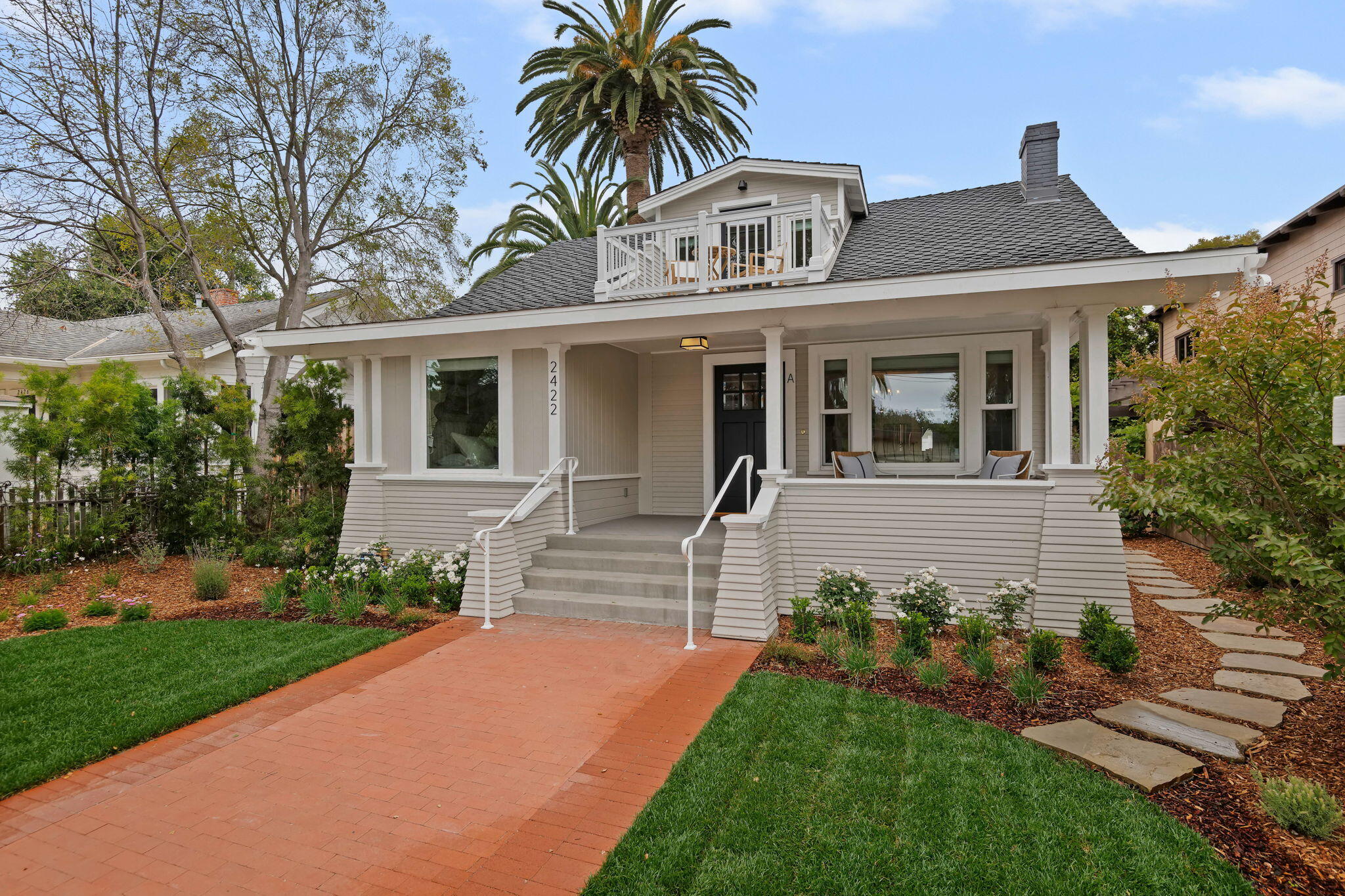 Light-colored house with a covered porch and upper balcony, approached by a brick walkway through landscaped gardens.