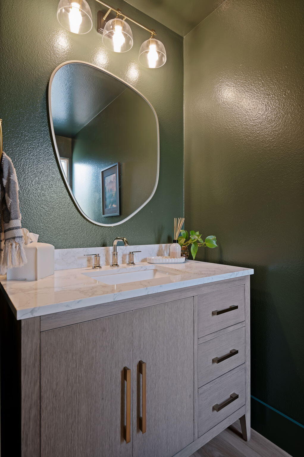 Bathroom vanity with marble countertop, gray wood cabinetry, oval mirror, and three pendant lights against dark green walls.