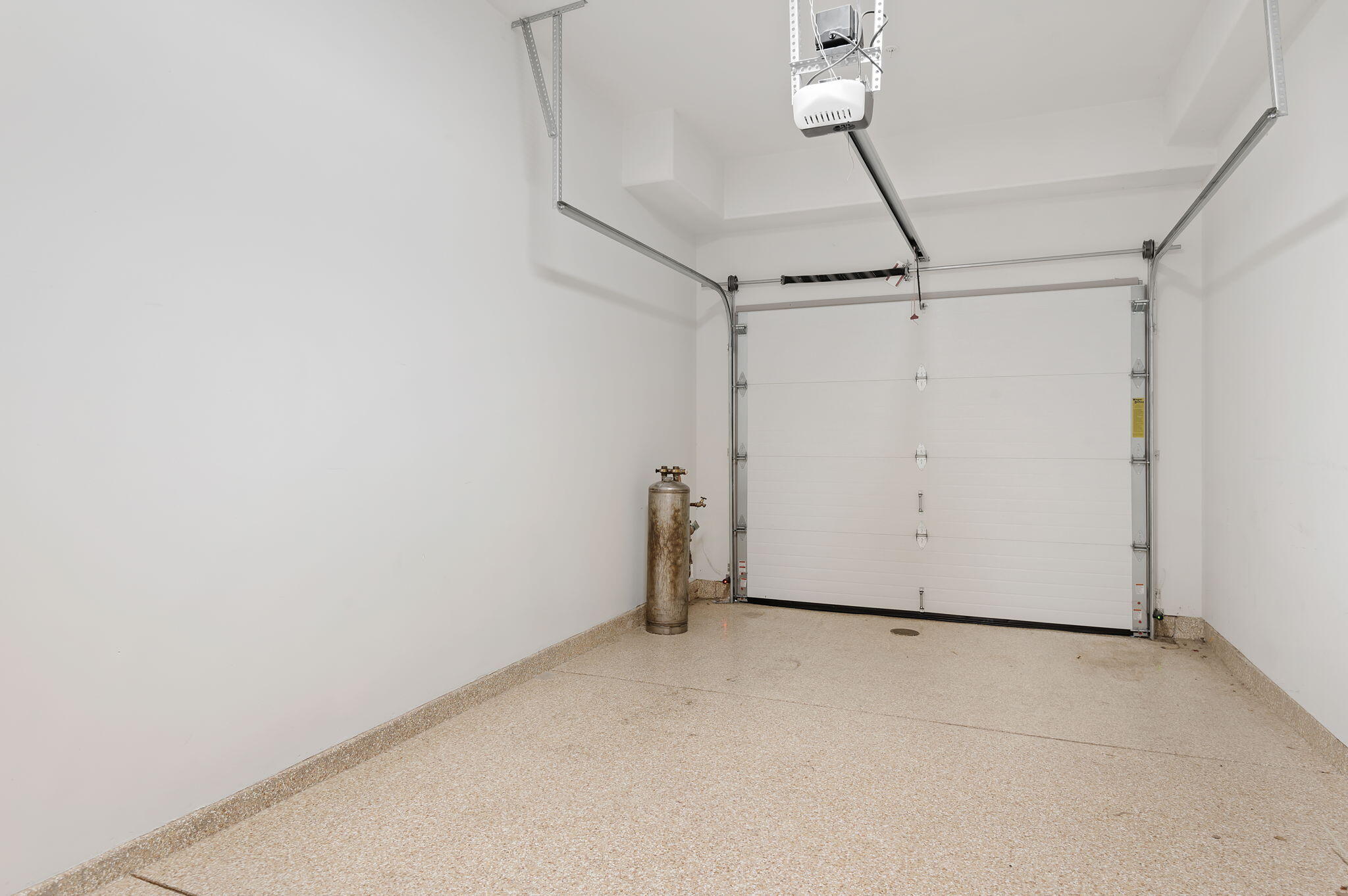 Empty garage with a closed overhead door, ceiling-mounted opener, and speckled concrete floor.