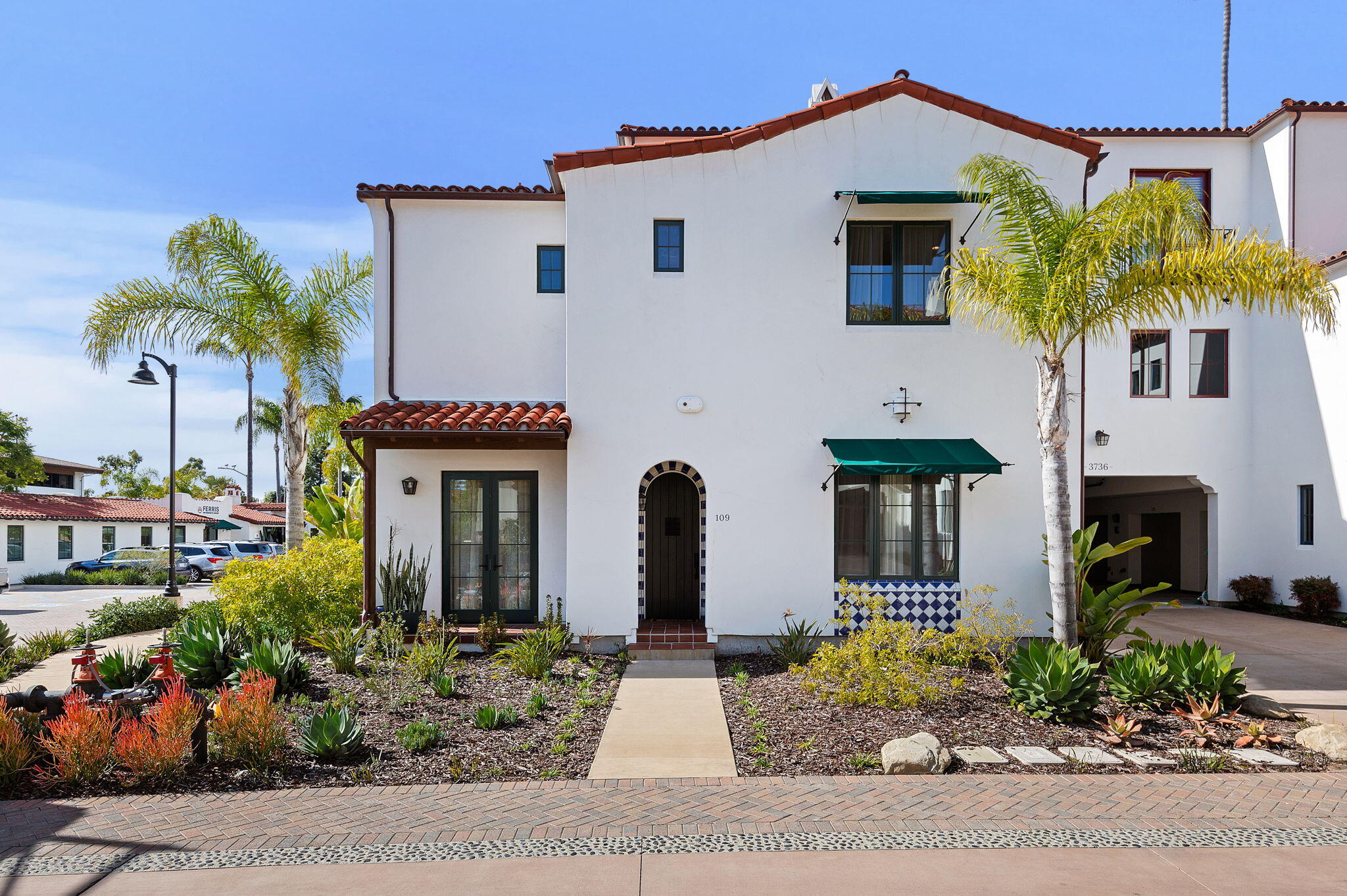 White stucco building with red tile roofs, green awnings, palm trees, and a landscaped front entry.