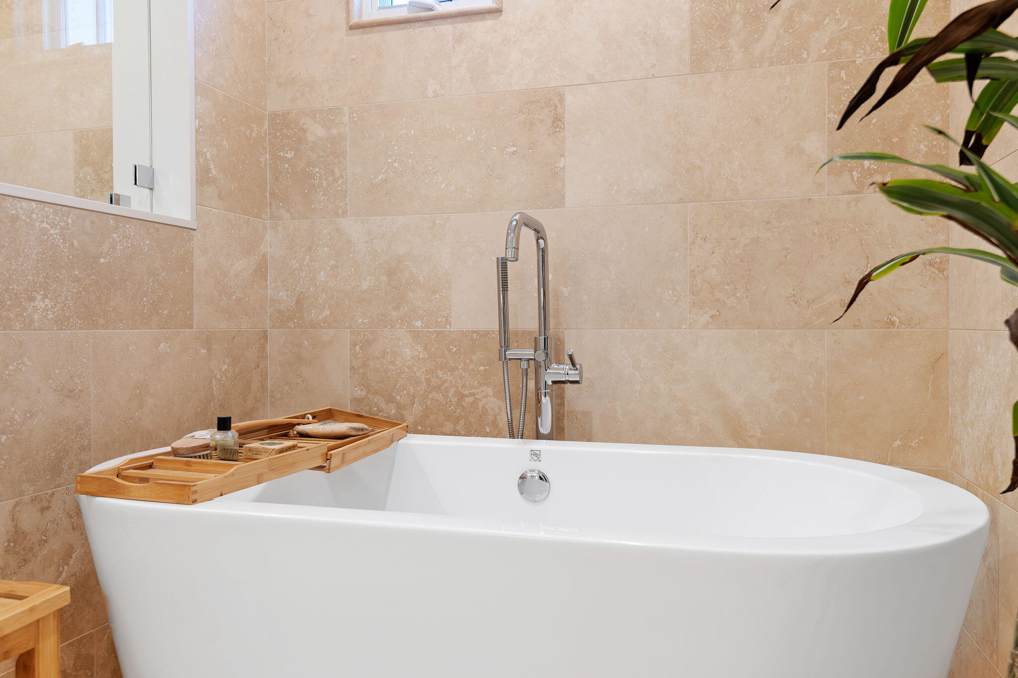 Freestanding white bathtub with chrome shower fixtures, a wooden bath tray, and tan stone-tile walls.