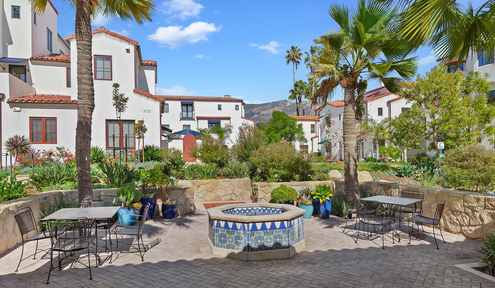 Residential courtyard with a tiled fountain, palm trees, patio seating, and mountains in the background.